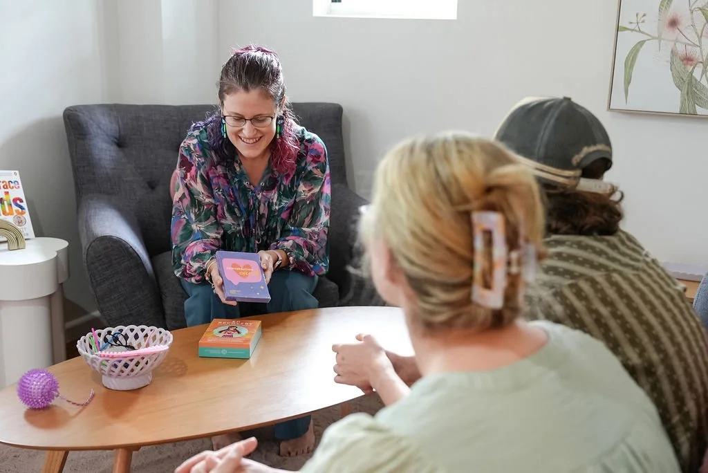A woman with glasses and dyed purple hair sitting on a gray armchair smiling while holding a tarot card reading deck, with two women seated at a wooden table in front of her.
