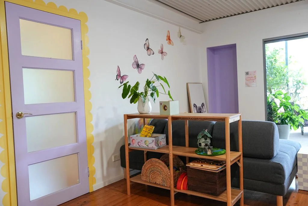 A living room with a purple door, a gray sofa, and a wooden shelf holding various decorative items. There's a plant on top of the shelf, butterfly wall decals on the white wall, and a large window with green plants outside.