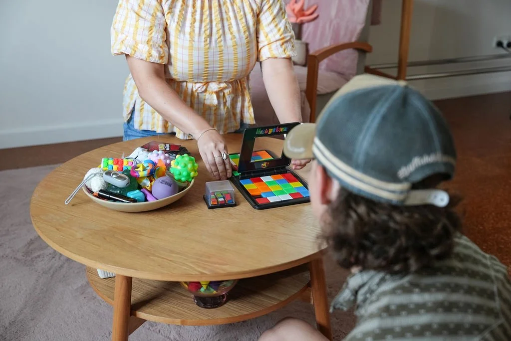 Child playing with colorful toys and makeup on a wooden table, while another person watches. The table has a bowl filled with various toys, and some colorful makeup palettes.