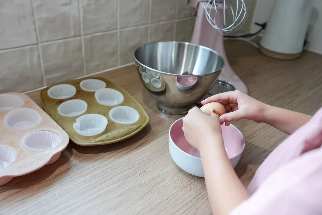 A person cracking an egg into a pink bowl in a kitchen with silicone cupcake molds, a large metal mixing bowl, and a pink stand mixer on a wooden counter.