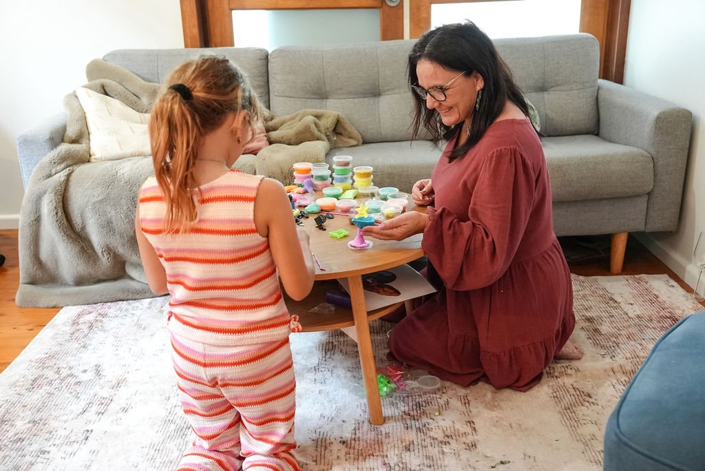 A woman and a girl playing with colorful clay on a small wooden table in a living room.