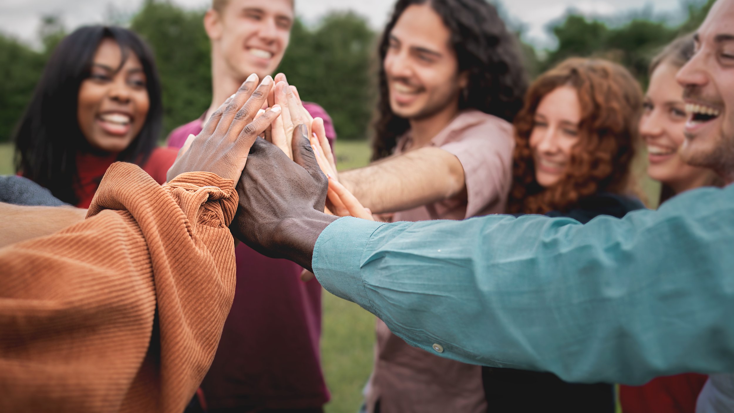 People of diverse ethnicities placing their hands together in a teamwork gesture outdoors, smiling and appearing happy.