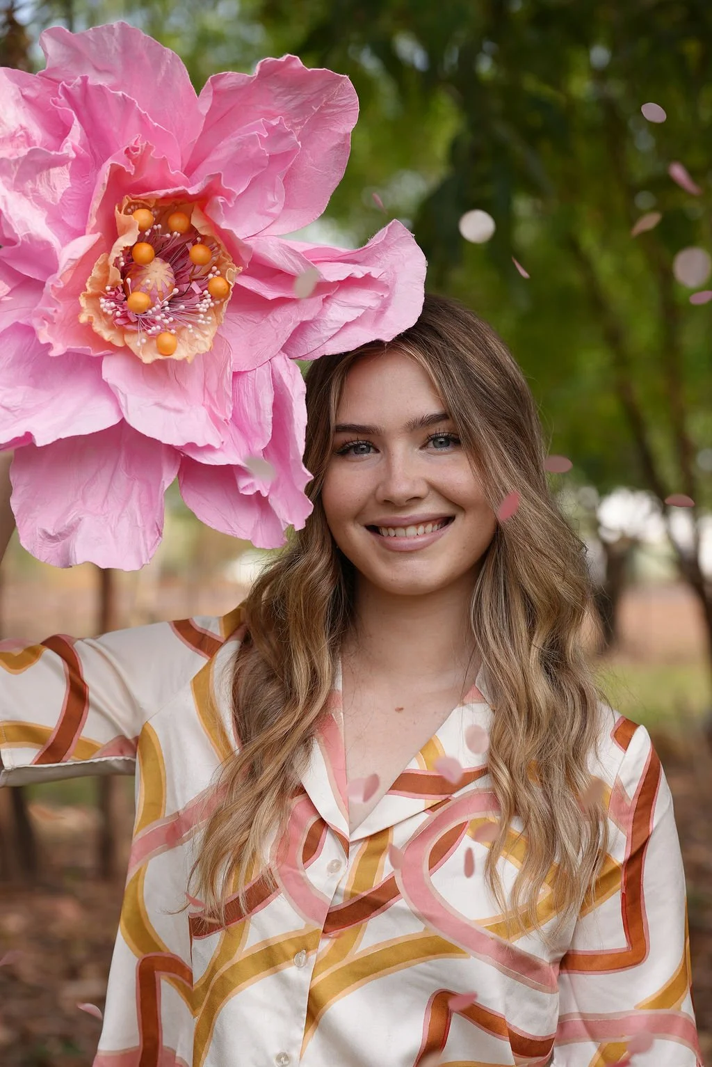 A smiling young woman with long wavy blonde hair in a patterned blouse, outdoors in a wooded area, with a large pink flower and falling pink petals near her head.