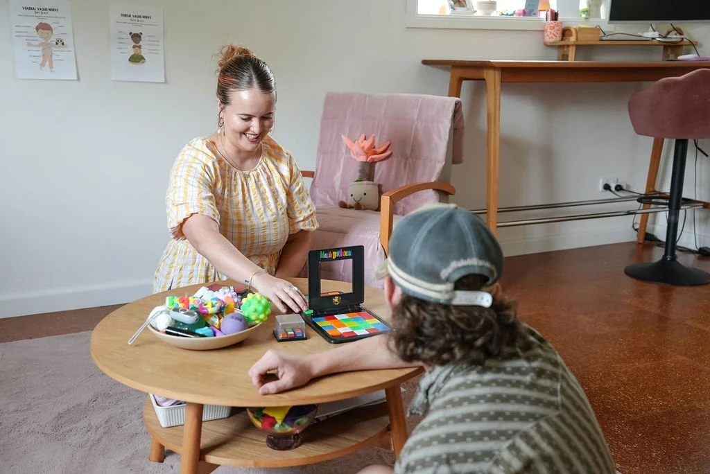 A woman and a child playing board games at a round wooden table in a living room. The woman is smiling and reaching for toys on the table, which include a colorful board game, a bowl of toys, and a Rubik's Cube. The woman has brown hair tied back, and the child wears a gray cap and striped shirt. The background shows a pink armchair with a decorative pillow, a wooden desk, and a window with various items on the windowsill.
