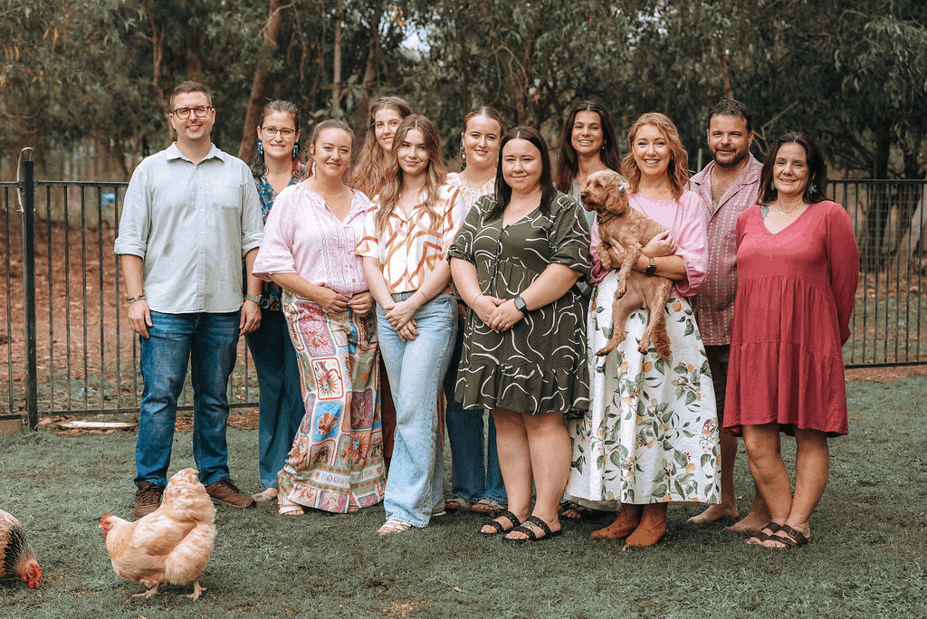 Group of flourish clinicians standing outdoors on grass, with a fence and trees in the background, some holding a dog and surrounded by chickens