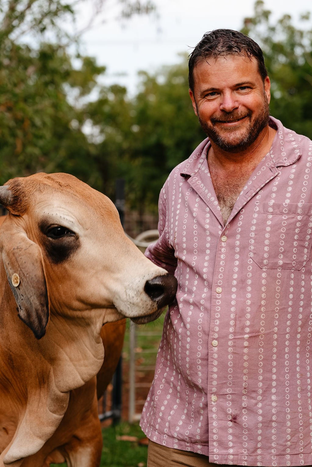 A man with dark hair and a beard smiling next to a brown and white cow in a grassy outdoor area.