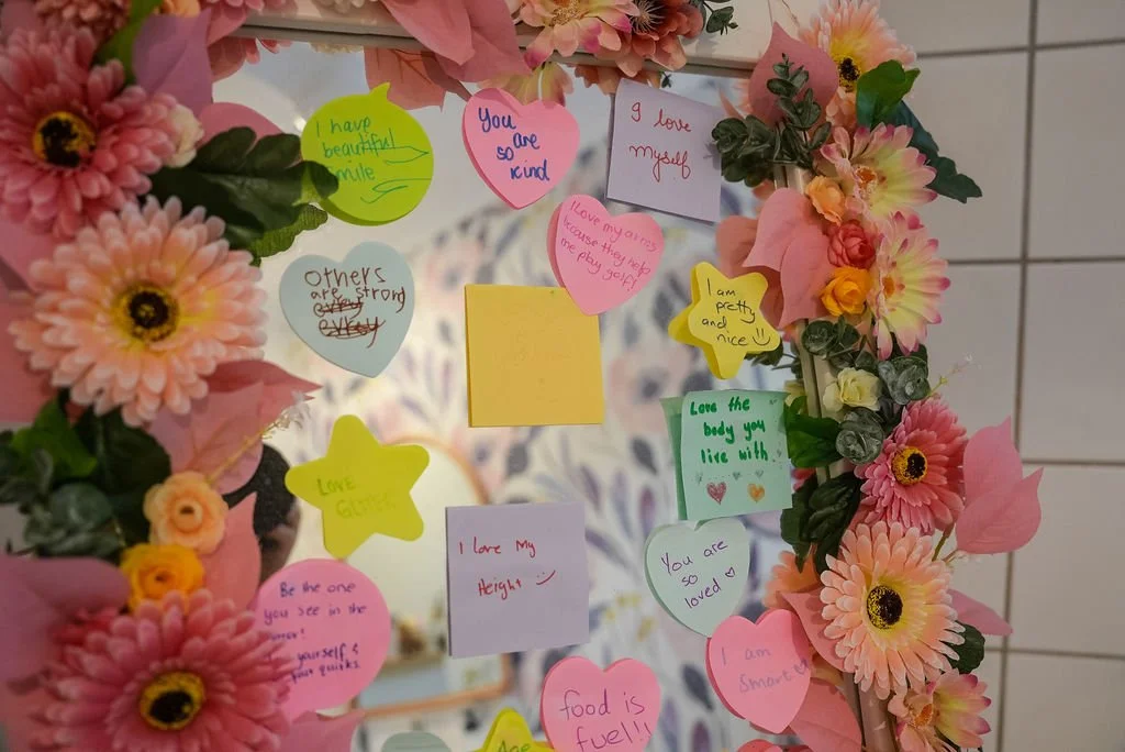 Decorative mirror with pink and yellow flowers around the frame, covered in colorful sticky notes with positive messages such as 'You are so kind,' 'I love myself,' and 'Love the body you like with!'