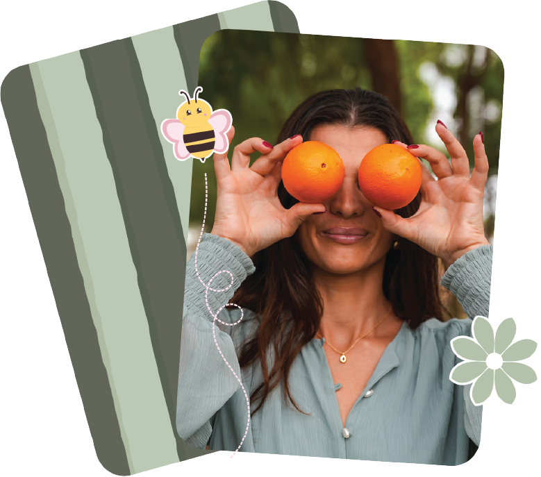 A woman holding two oranges in front of her eyes, smiling outdoors.