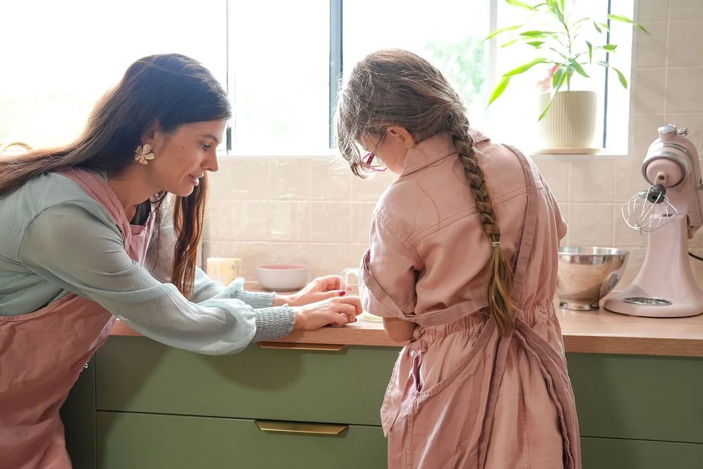 A woman and a young girl baking together in a bright kitchen, with a stand mixer and potted plant on the counter.