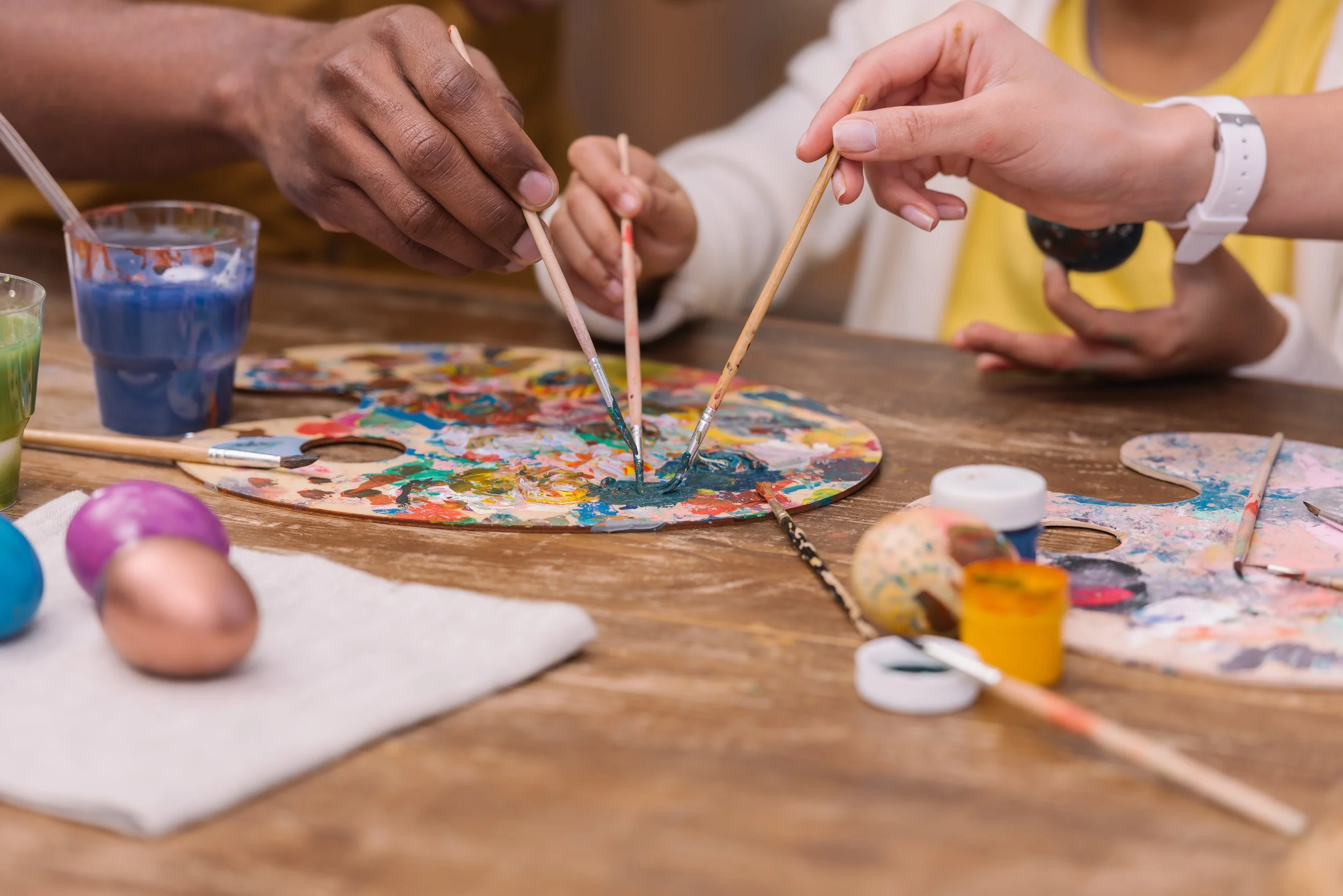 People painting Easter eggs with paintbrushes on a wooden table with paint and art supplies.