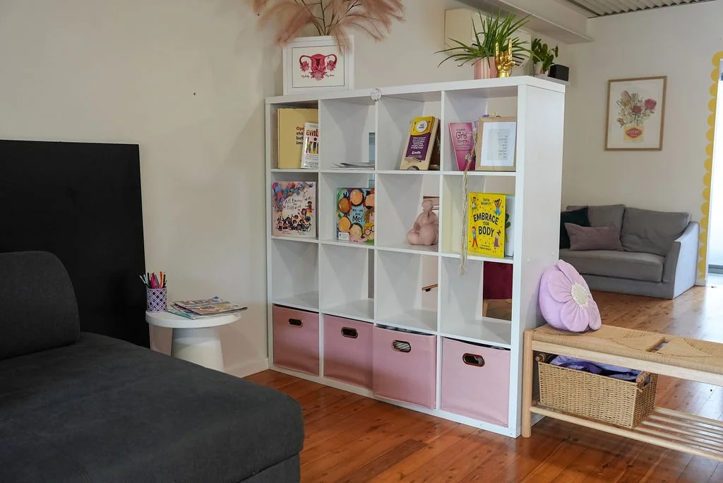 A white bookshelf with pink storage bins at the bottom, decorated with books, plants, and decorative items, in a cozy living room with wooden floors and a gray couch.