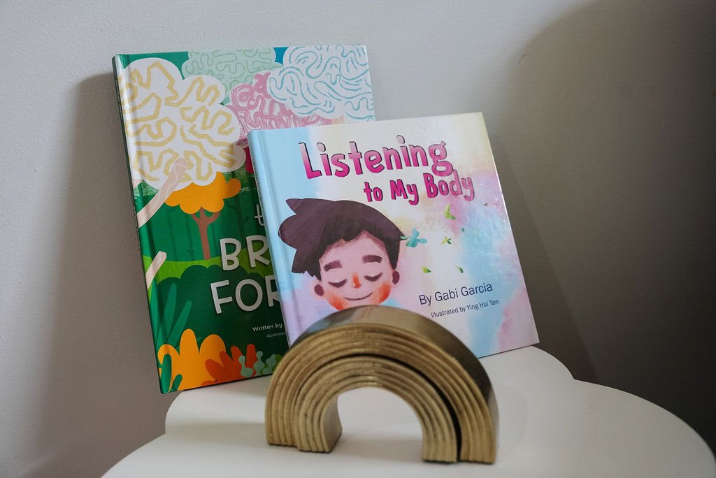 Two children's books, one titled "Listening to My Body" by Gabi Garcia and the other with a colorful abstract cover, leaning against a wall on a white surface, with a wooden rainbow stacker in front.