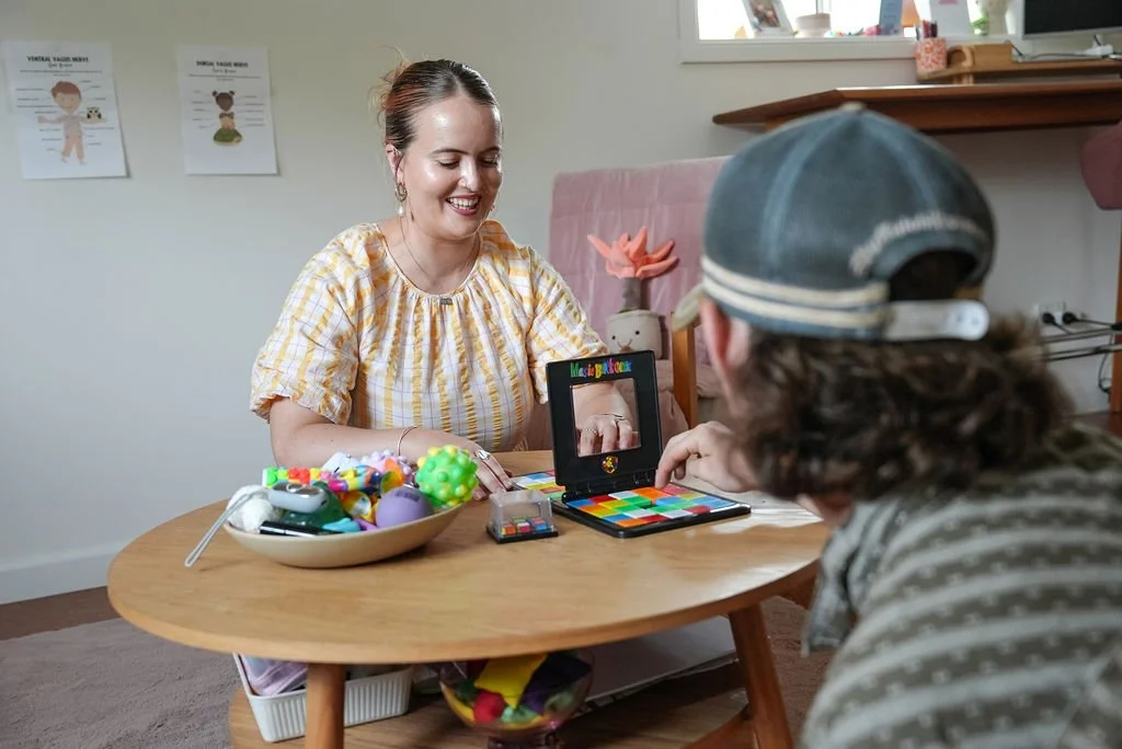 A woman and a child sitting across from each other at a small wooden table playing a colorful game. The woman is smiling, wearing a yellow checkered top, and has earrings. The child, wearing a hat and striped shirt, is pointing at the game. There is a bowl of toys on the table, and posters on the wall behind them.