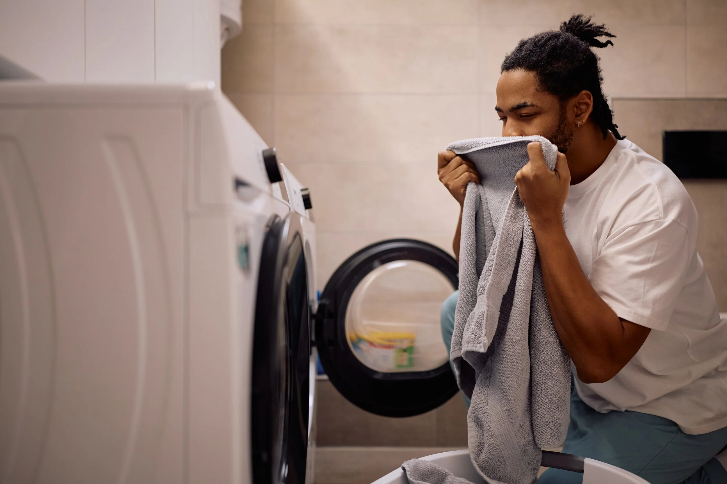 A man with dreadlocks smells a gray towel in front of a washing machine in a laundry room.