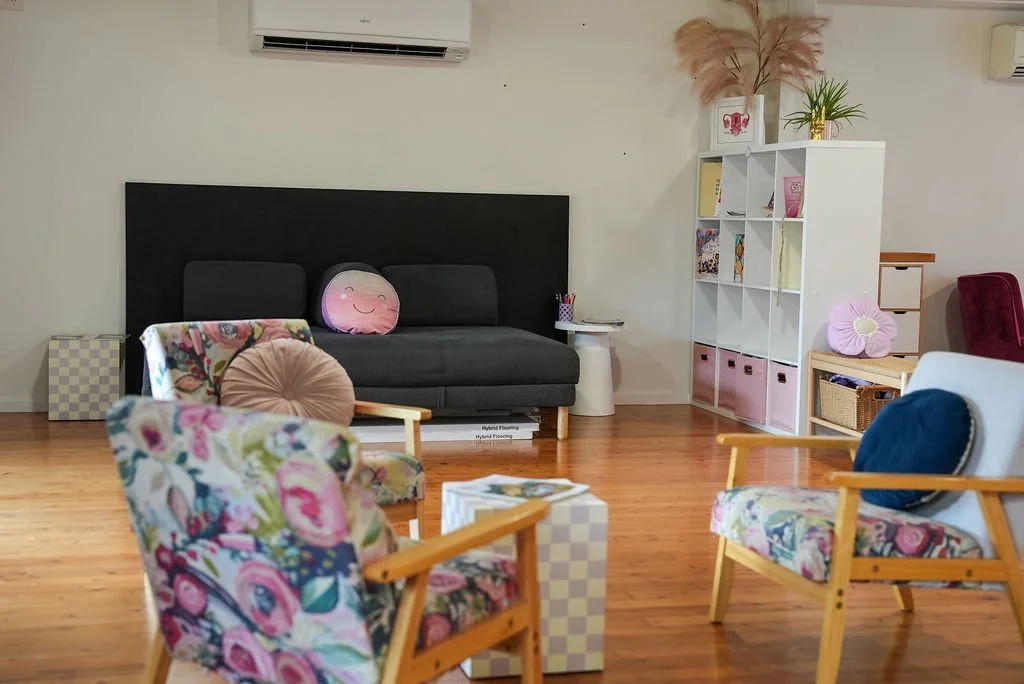 Living room with wooden floors, floral armchairs with colorful cushions, a black couch with a pink round pillow, a tall white shelf, and decorative plants.