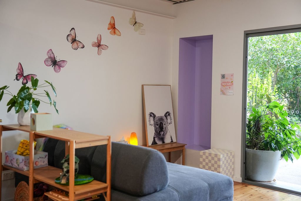 A cozy living room with butterfly wall decals, a painting of a koala, and a sliding door leading outside to greenery.