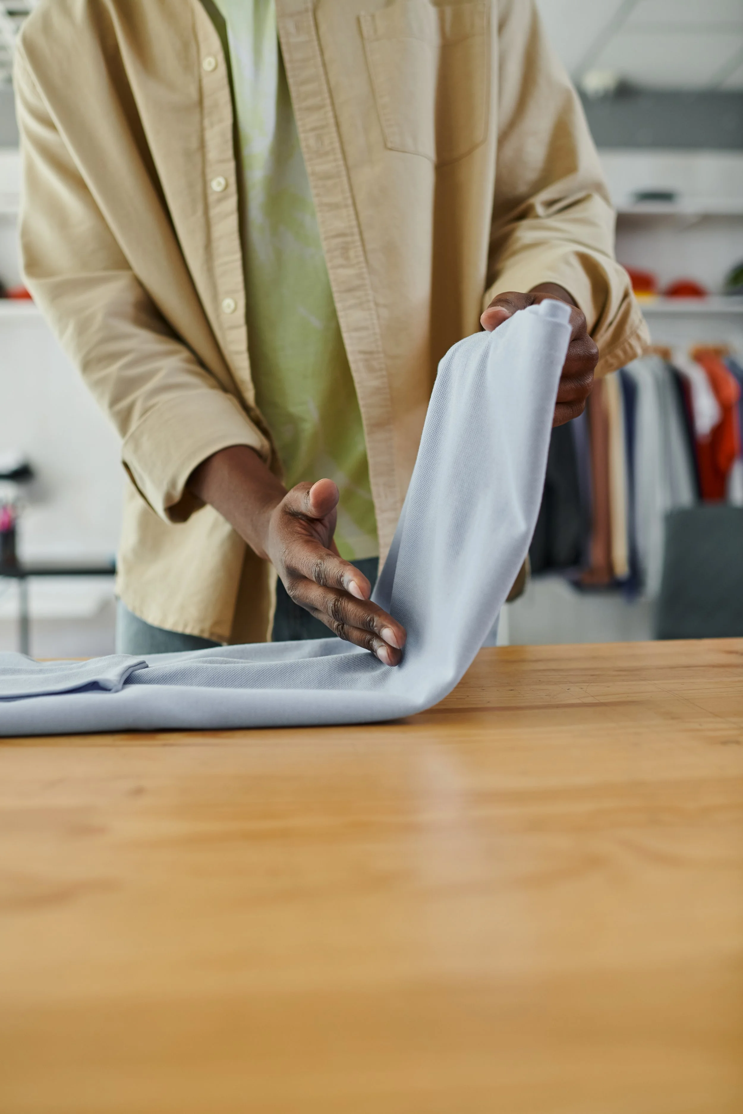 Person folding a light gray fabric or clothing item on a wooden table in a retail or clothing store environment.