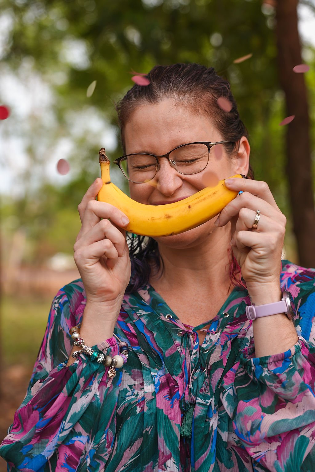 Woman with glasses and colorful shirt smiling with eyes closed, holding a banana in front of her face as if it were a smile, with falling pink and white flower petals in the background.