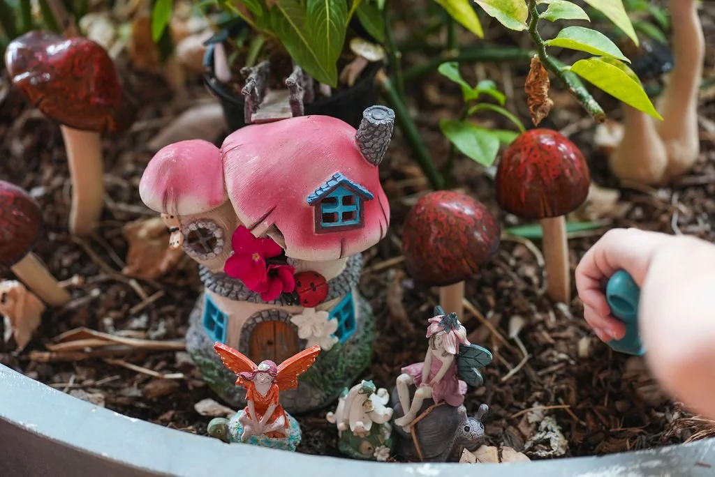 A fairy tale-themed miniature garden with mushroom and fairy figurines, including a pink mushroom house, surrounded by red mushrooms, greenery, and small decorative fairy characters.