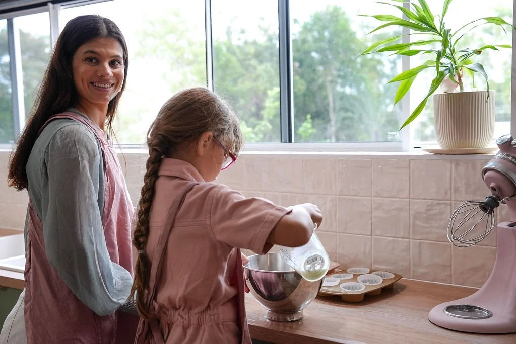 A woman and a young girl baking together in a kitchen. The girl is pouring ingredients into a mixing bowl while the woman smiles at the camera. The kitchen has a large window, a potted plant, and a stand mixer on the counter.