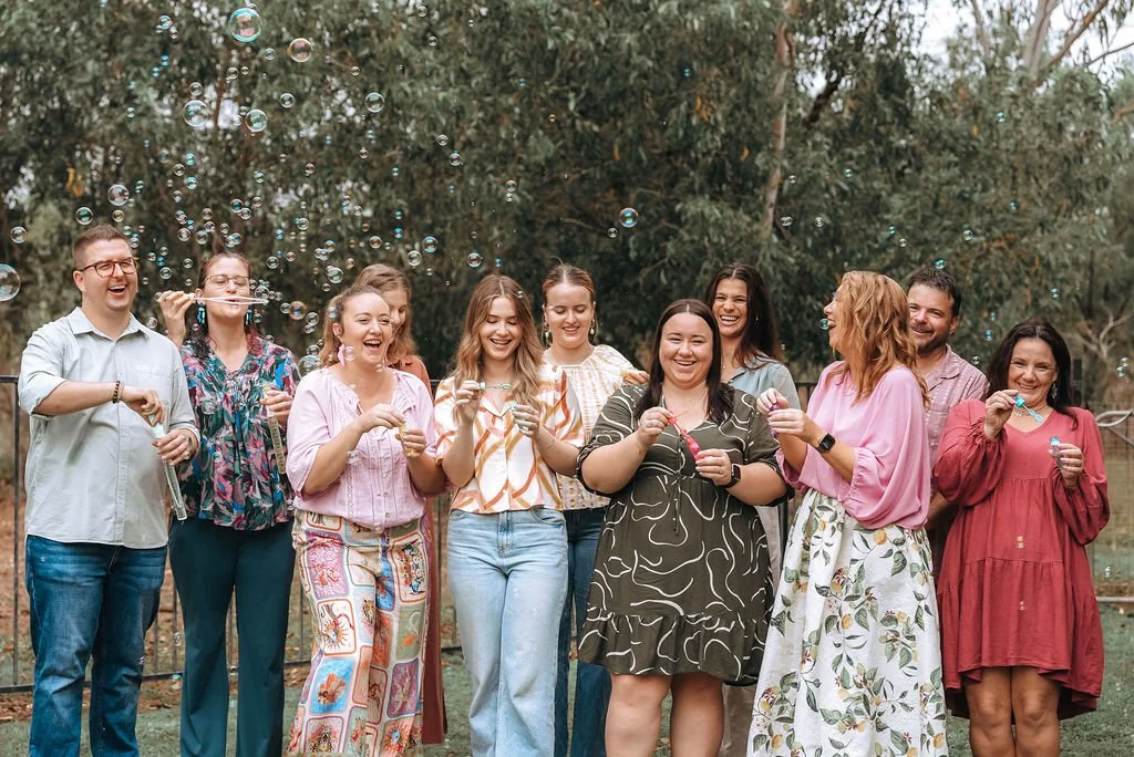 Group of people outdoors, blowing bubbles and laughing, with trees in the background.