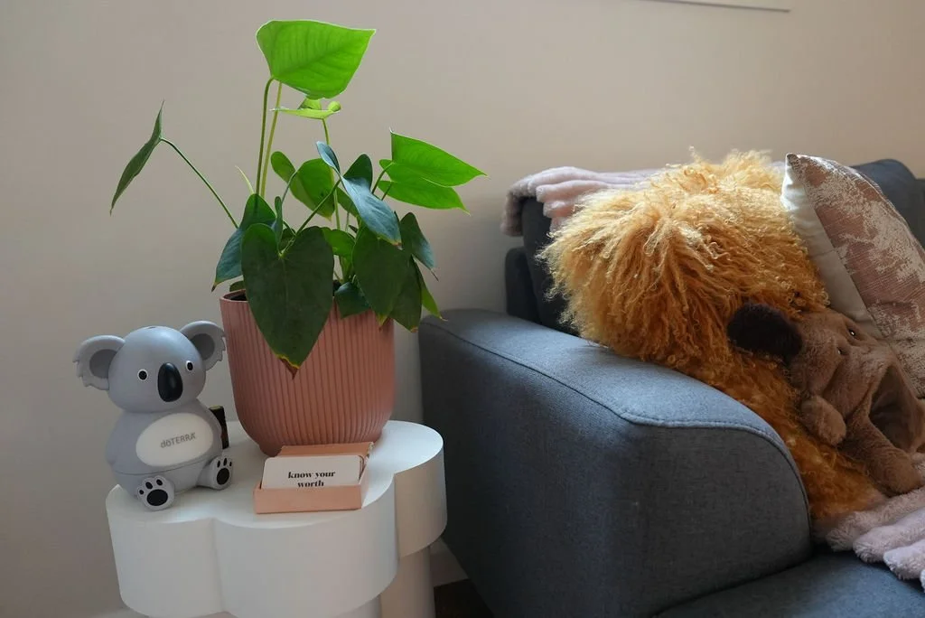 A therapy room with a plant and koala on a small table, comfy grey coach with a mustard throw pillow.