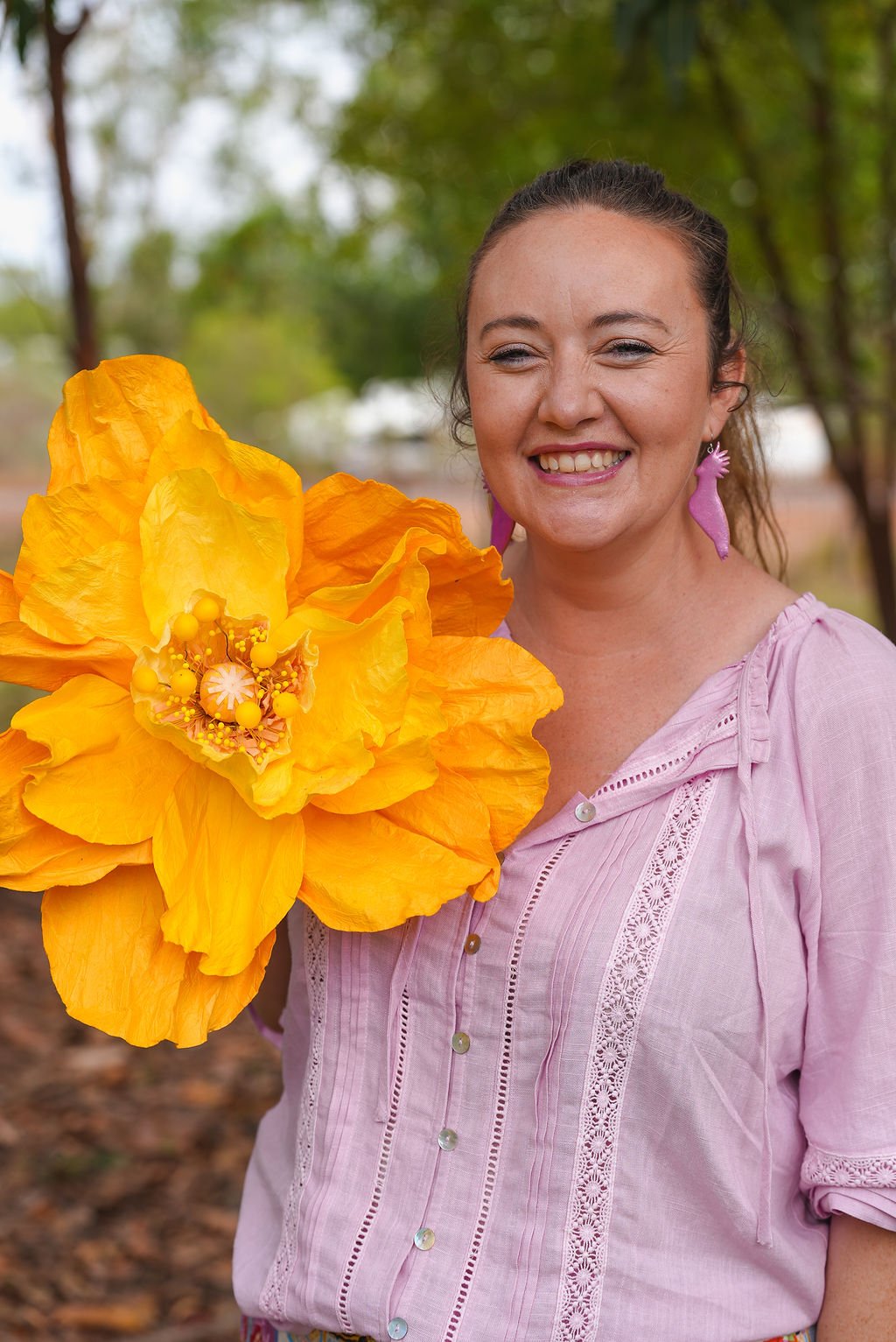 A woman smiling outdoors, holding large orange and yellow artificial flower, wearing a pink top with purple earrings shaped like seahorses, with green trees in the background.