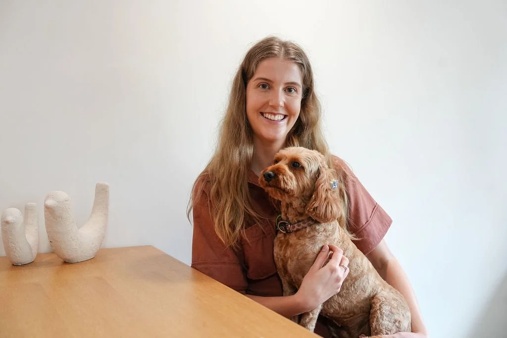 A smiling young woman with long wavy hair sitting at a wooden table, holding a tan-colored dog with curly fur in front of a plain white wall. There are three white ceramic vases on the table to her left.