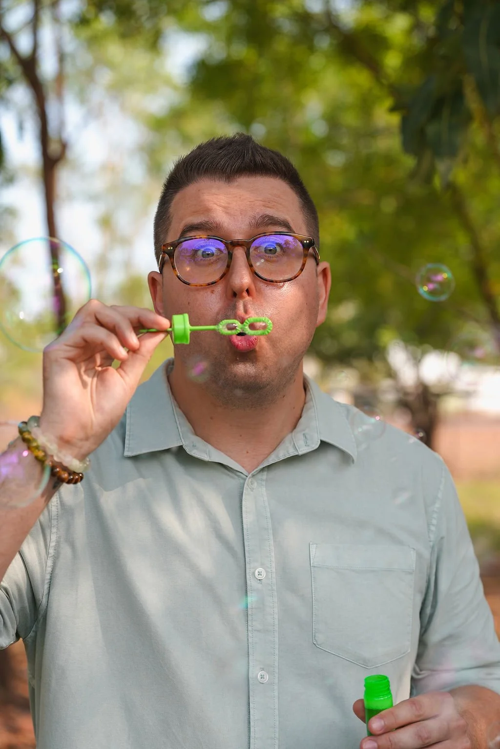 A man with glasses blowing bubbles outdoors, holding a bubble wand and a container of bubble solution, with a blurred green background of trees.