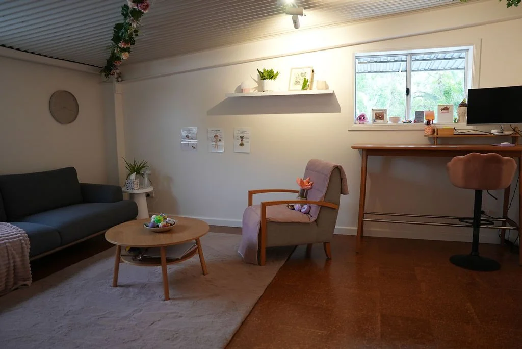 A cozy living room with a dark gray sofa, a round wooden coffee table holding a decorative bowl, a beige armchair with a pink throw blanket and flower pillow, a white wall shelf with decorative items, a window with a view of trees, and a workspace ar
