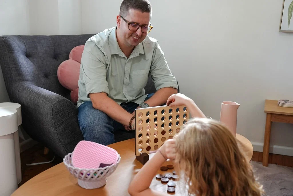 Clinician, Daniel, sits at a table with a young girl playing Connect Four.