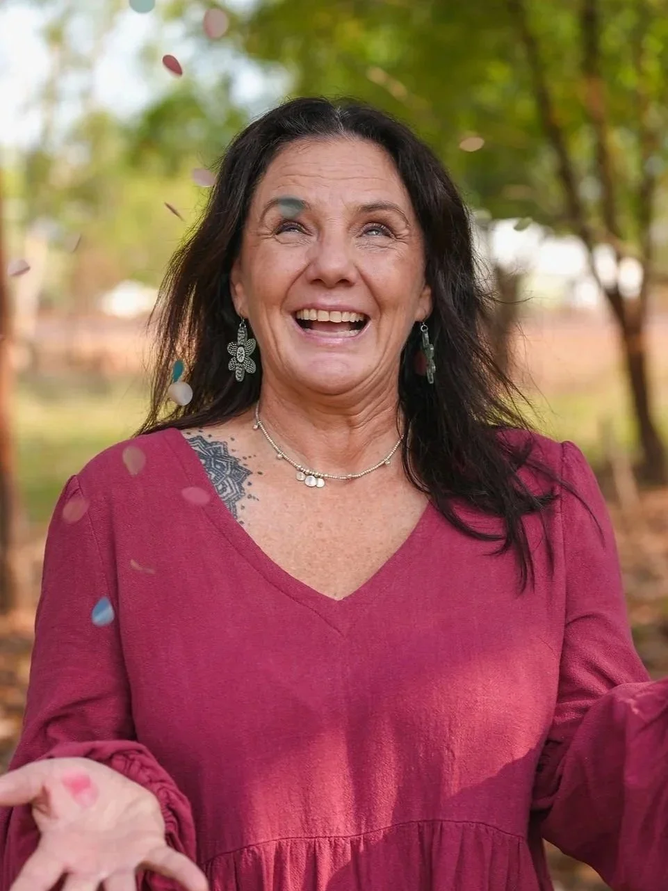 A woman with dark hair tied up, wearing earrings and an orange sleeveless top, smiling outdoors against a blurred natural background.