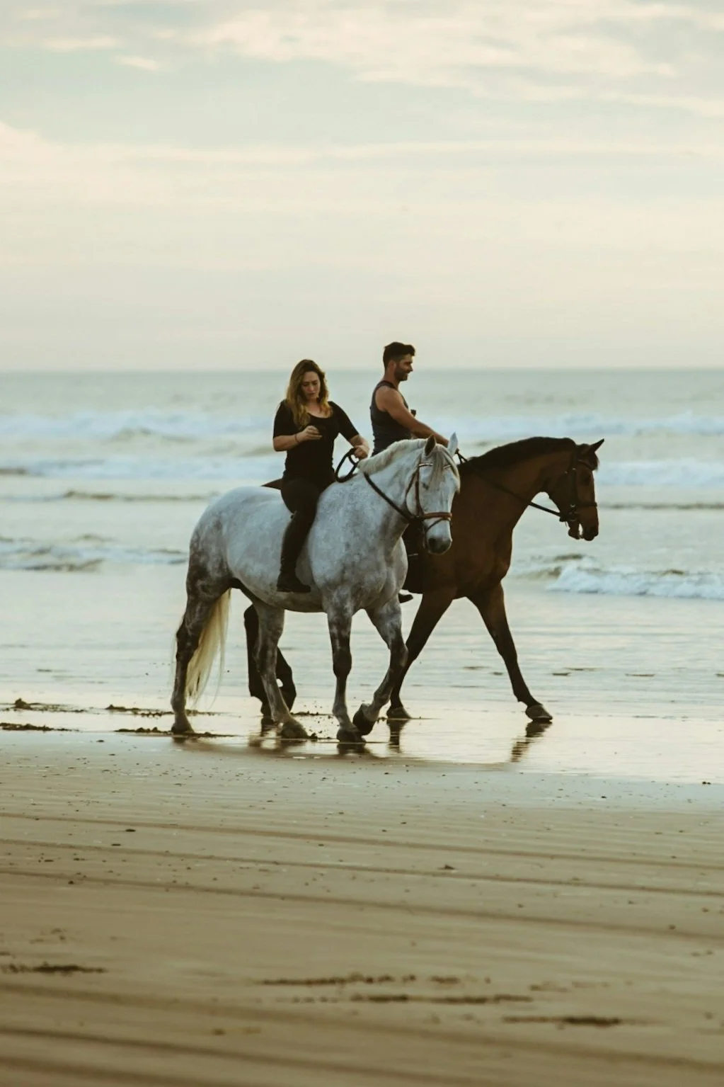 A woman and a man riding horses along the beach at sunset, with waves and cloudy sky in the background.