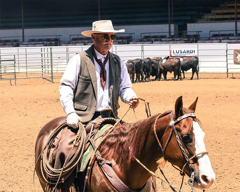 A man in cowboy attire, including a wide-brimmed hat, sunglasses, and vest, rides a horse inside an arena with a sandy surface.
