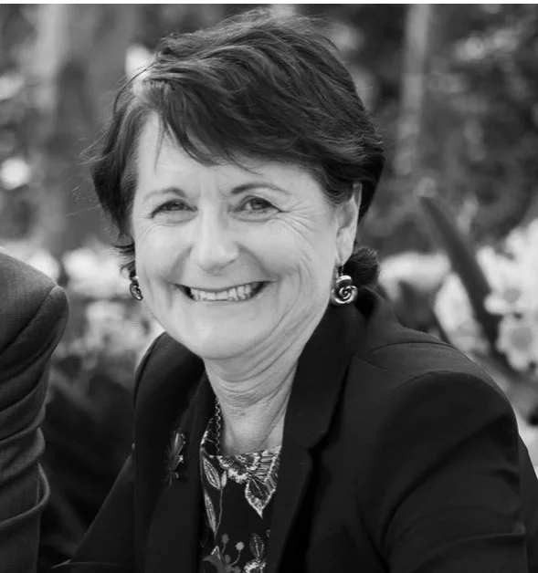 Black and white photo of a smiling woman with short hair, wearing earrings and a blazer, outdoors with trees in the background.