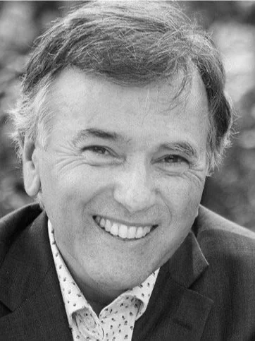 Black and white photo of a smiling man in a suit and patterned shirt.