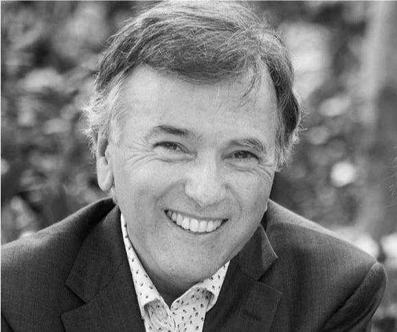 Black and white photo of a smiling man with short, light-colored hair, wearing a suit and a patterned shirt, outdoors.