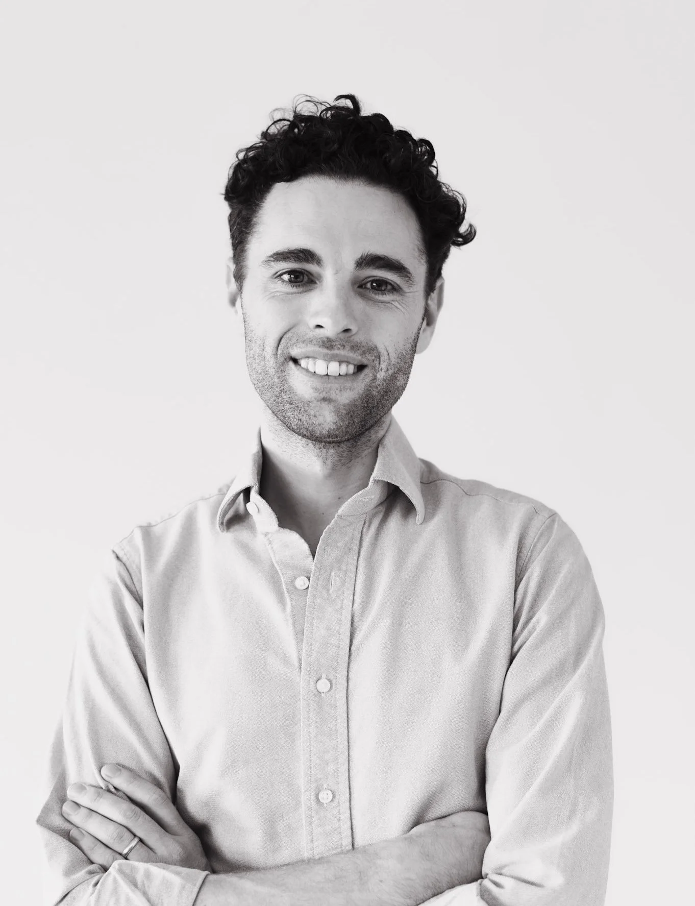 Black and white portrait of a smiling man with dark curly hair, wearing a light-colored button-up shirt with arms crossed.