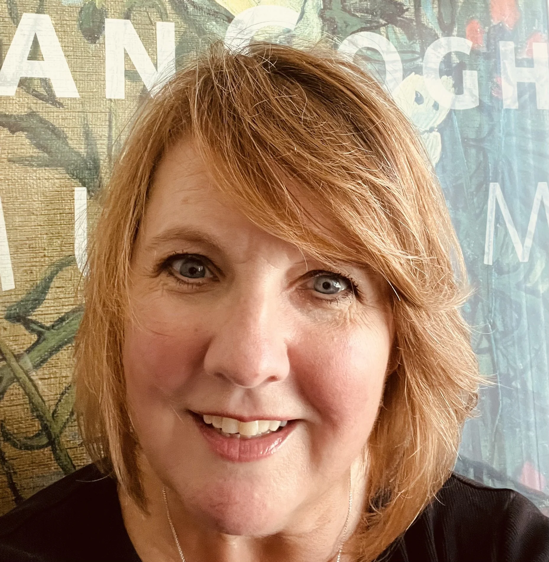 A woman with shoulder-length red hair and blue eyes smiling in front of a wall with faded text and floral patrones.