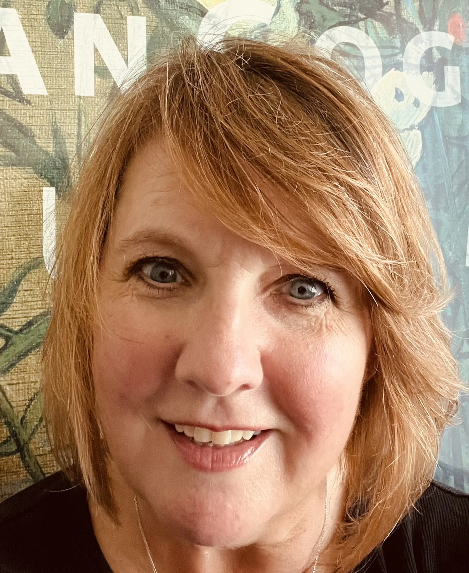 Close-up of a woman with short red hair, blue eyes, and a friendly smile, with a colorful background featuring text and floral patterns.