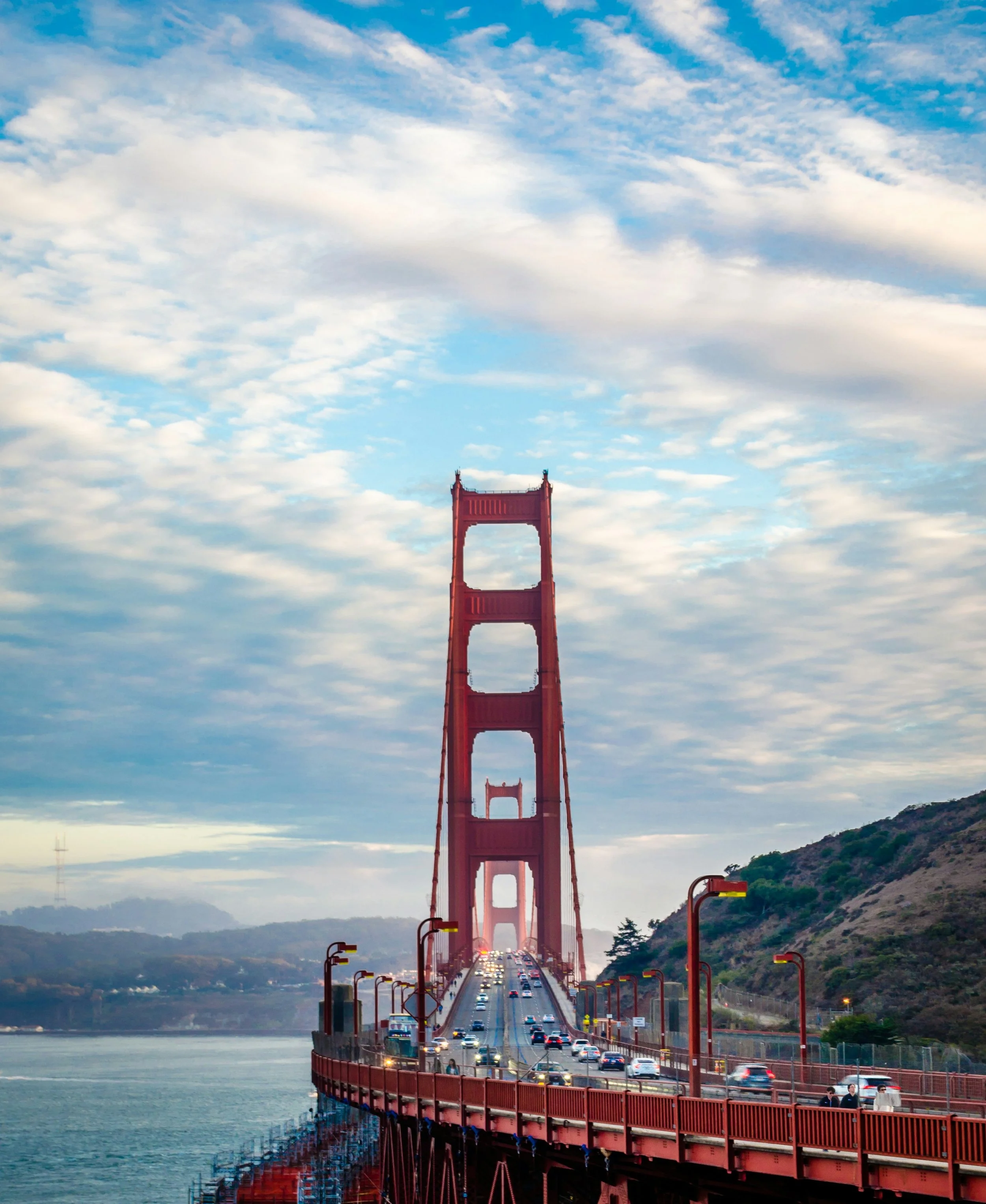 A view of the Golden Gate Bridge with cars moving across it, set against a partly cloudy sky with mountains in the background.