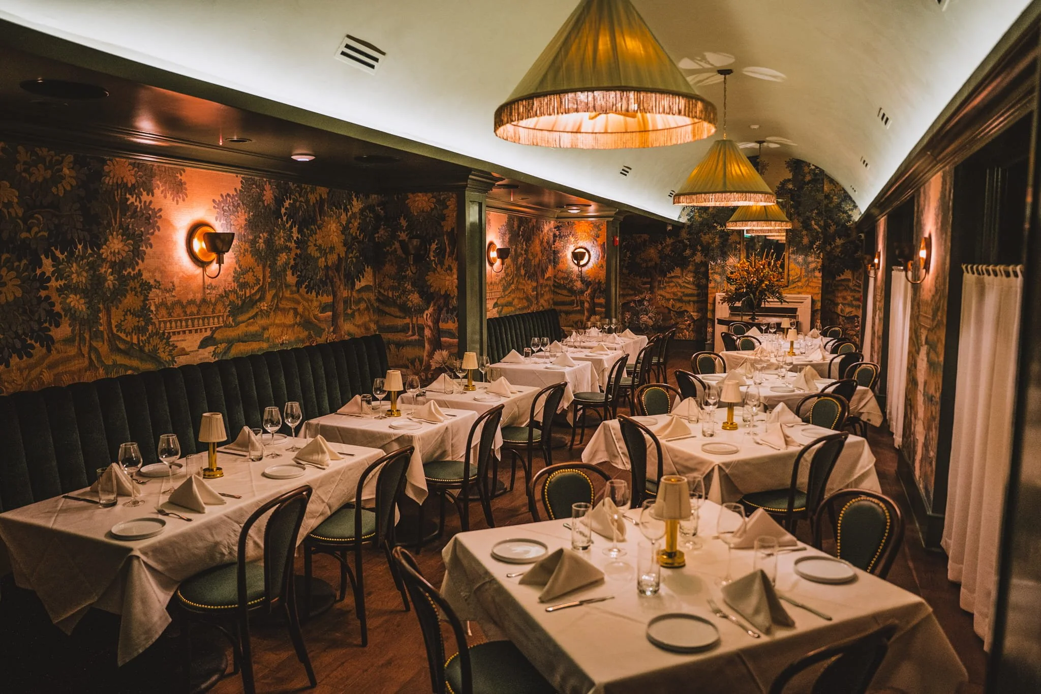 Elegant restaurant interior with tables set with white tablecloths, napkins, glassware, and small lamps, decorated with floral wallpaper, large green upholstered chairs, and hanging lamps, creating a warm and sophisticated atmosphere.