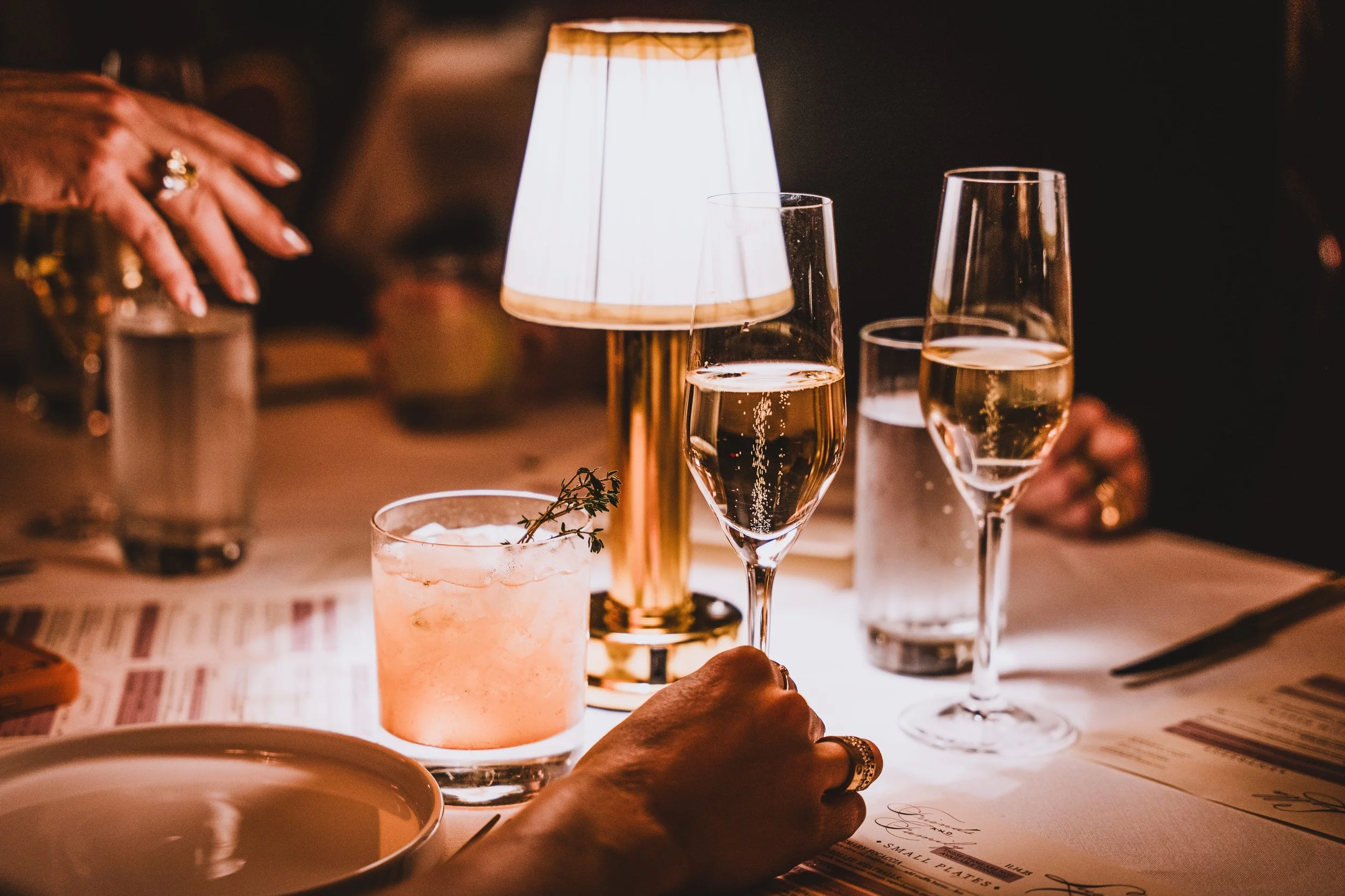 Close-up of a dimly lit table with various glasses of champagne and a pink cocktail, a small lamp emitting warm light, and a hand with rings reaching for a glass, with a menu on the table.