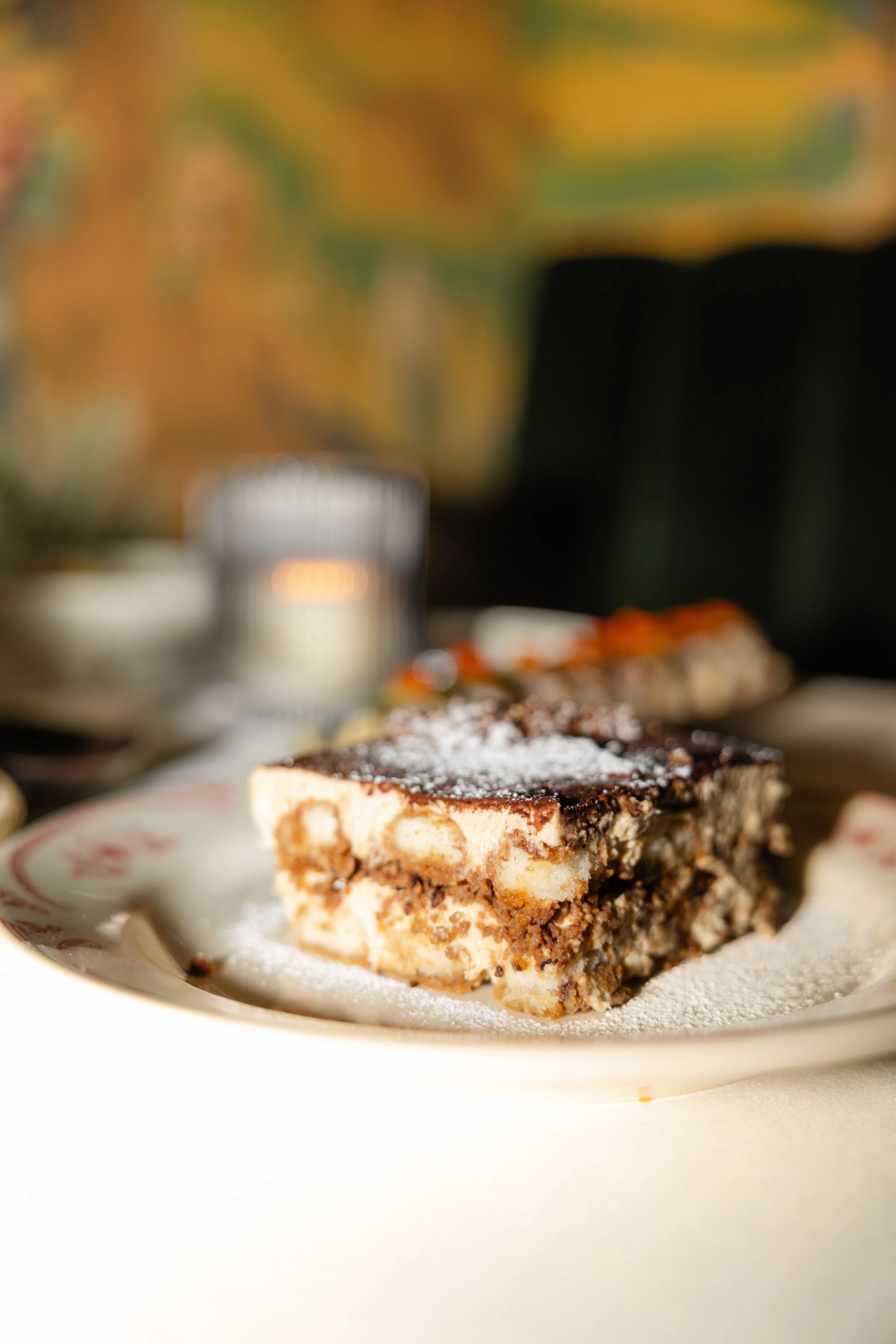 A close-up of a layered dessert with chocolate and vanilla layers, topped with powdered sugar, served on a decorative plate with a red pattern. In the blurred background, there is a glass of milk and some other food items, with a colorful autumn-them