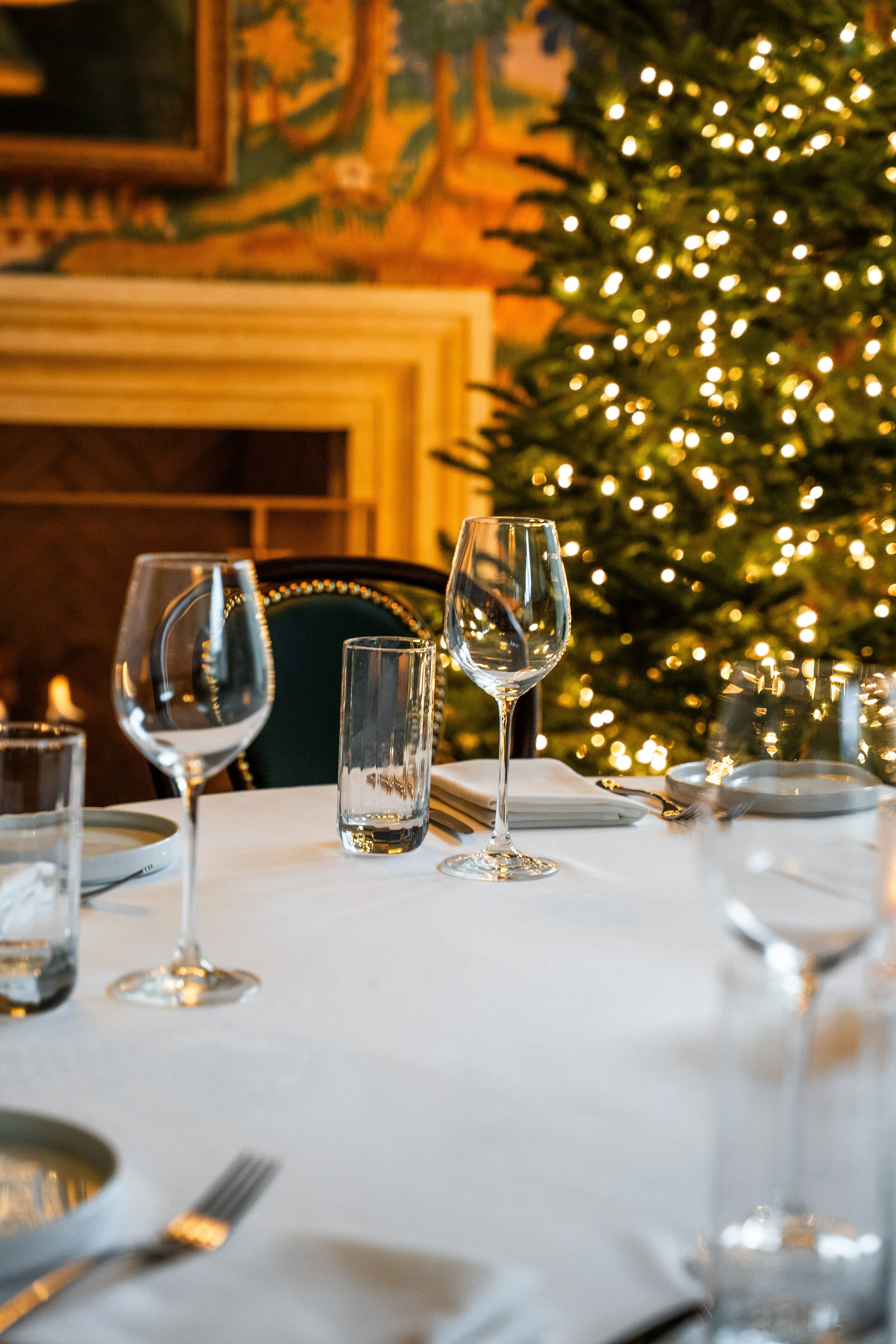 A table set with glassware, napkins, and utensils in front of a decorated Christmas tree with lights, with a mural painting and a fireplace in the background.