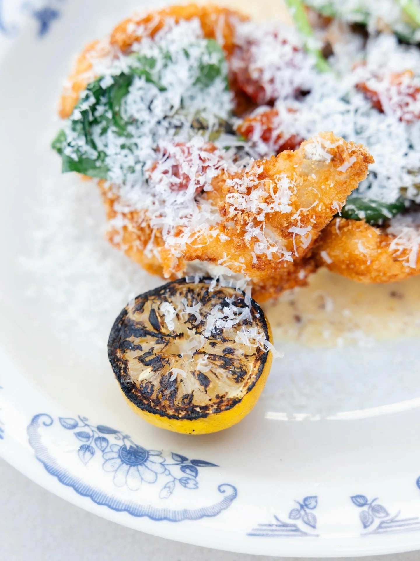 Close-up of grilled lemon half and fried food topped with shredded cheese and herbs on a white plate with a blue floral pattern.