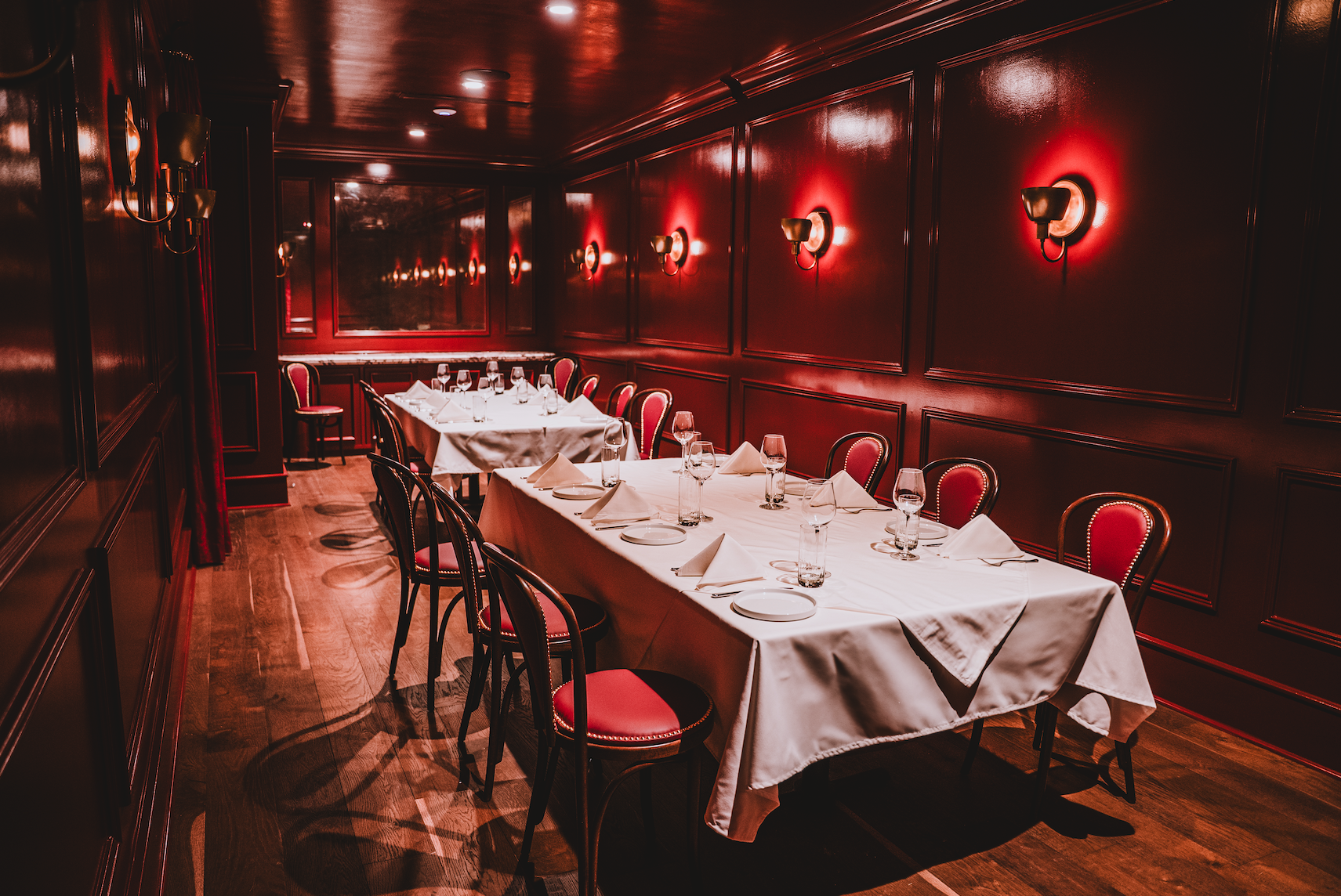 A private dining room with a long table set for a meal, featuring white tablecloths, napkins, wine glasses, water glasses, white plates, and cutlery, with red upholstered chairs against dark red paneled walls illuminated by wall sconces.