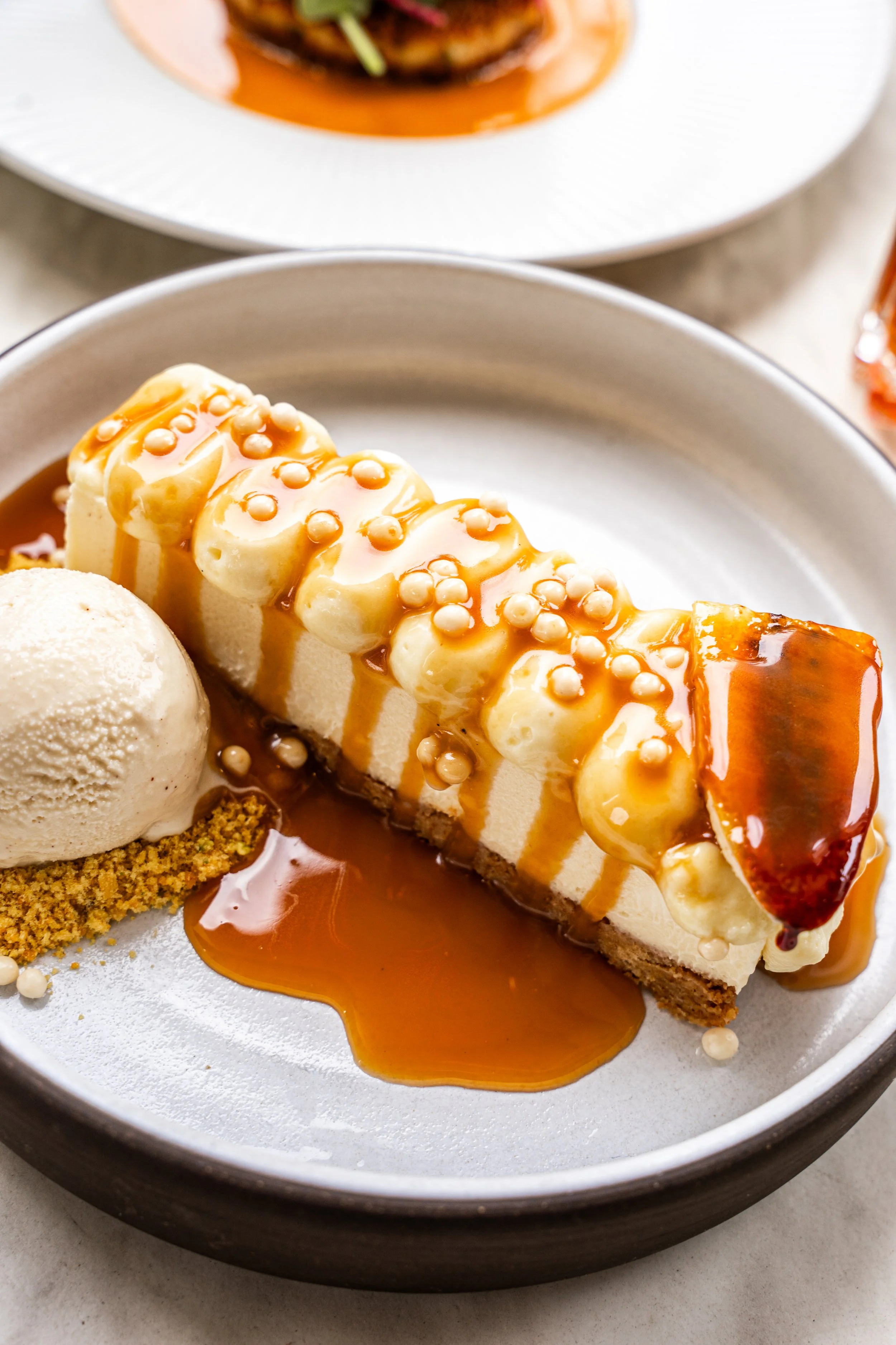 A slice of cheesecake topped with caramel sauce, banana slices, and decorative white sprinkles, served with a scoop of vanilla ice cream on a white plate.