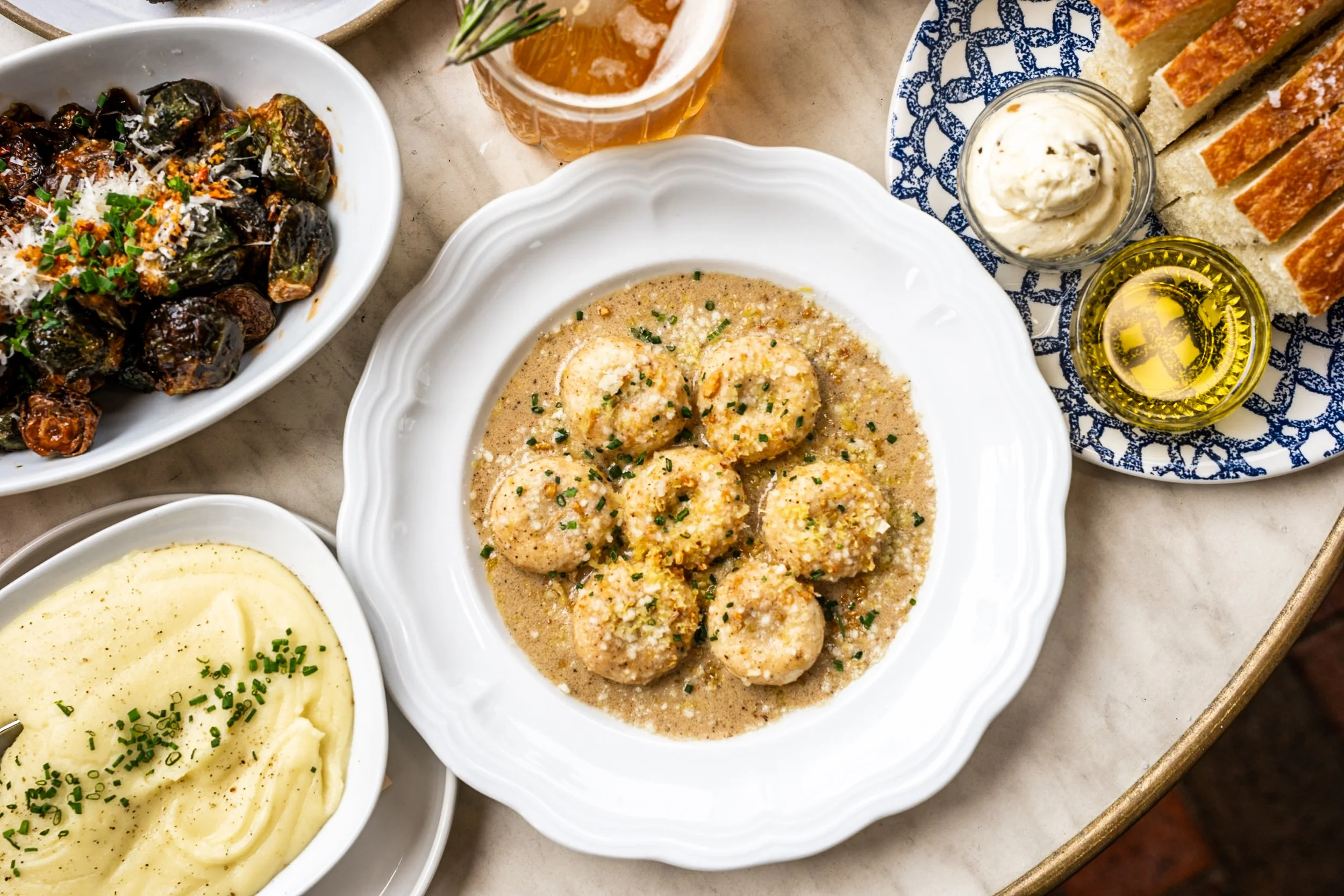 Assorted dishes on a table including mashed potatoes, a plate of meatballs in gravy, a bowl of roasted vegetables topped with cheese, and slices of bread with a cream spread. Also visible are a glass of beer and small jars of oil and butter.