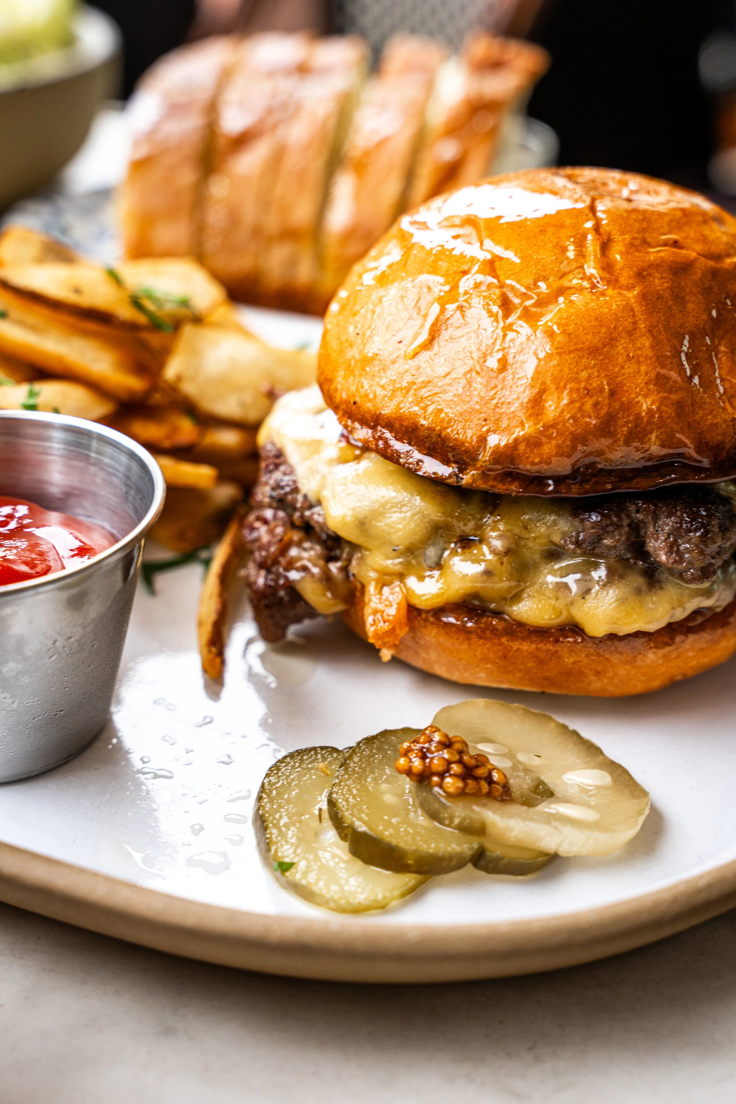 A cheeseburger with a bun, melted cheese, beef patty, and cheese sauce, served on a white plate with pickles, potato wedges, and ketchup.